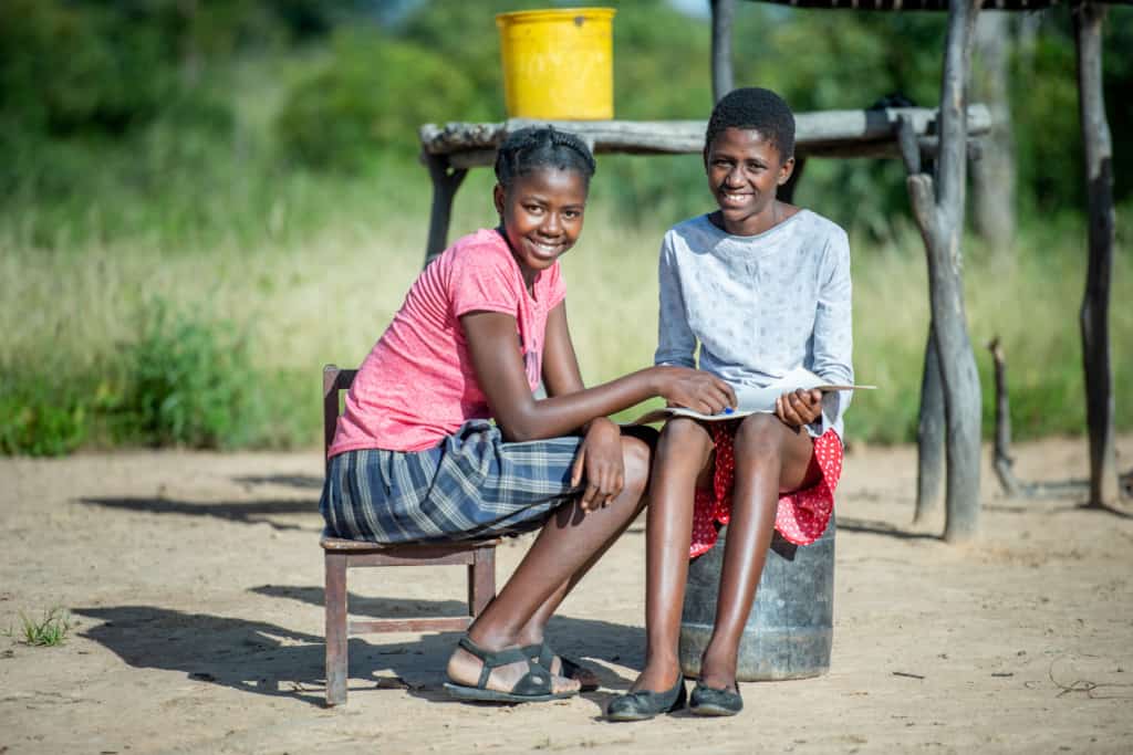 Two young African girls sit on chairs next to a water pump.