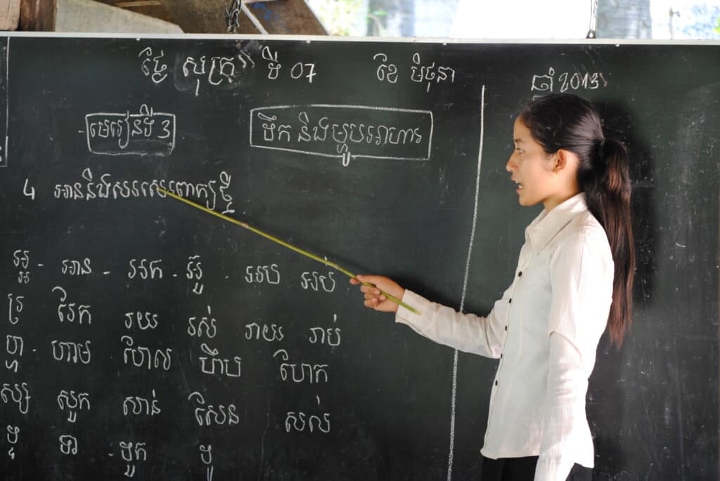 Teacher in Cambodia stands in profile in front right of a chalk board, uses a pointer to point to Khmer words written on the board