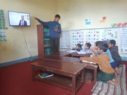 Teacher in Nepal points to TV screen at the front of the classroom while group of students sitting around tables look on 