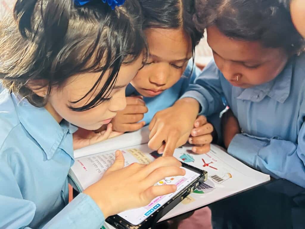 3 young girls gather around a phone resting on top of a book and do learning activities