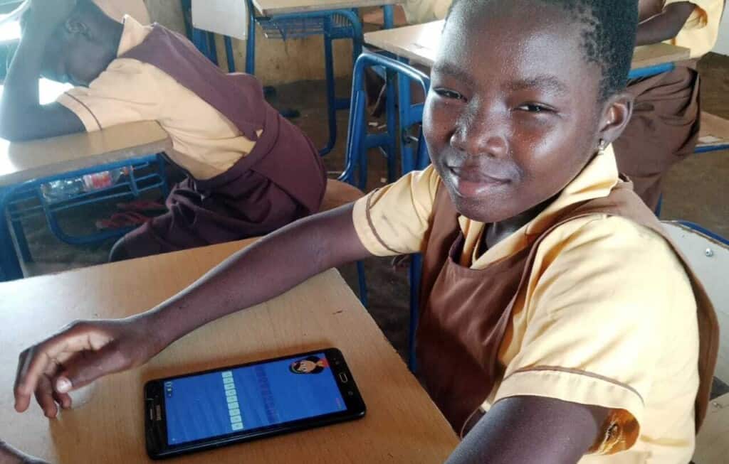 Young girl in Ghana sits at a desk in a classroom in her school uniform with a smartphone on the desk in front of her with the Google Read Along app on it