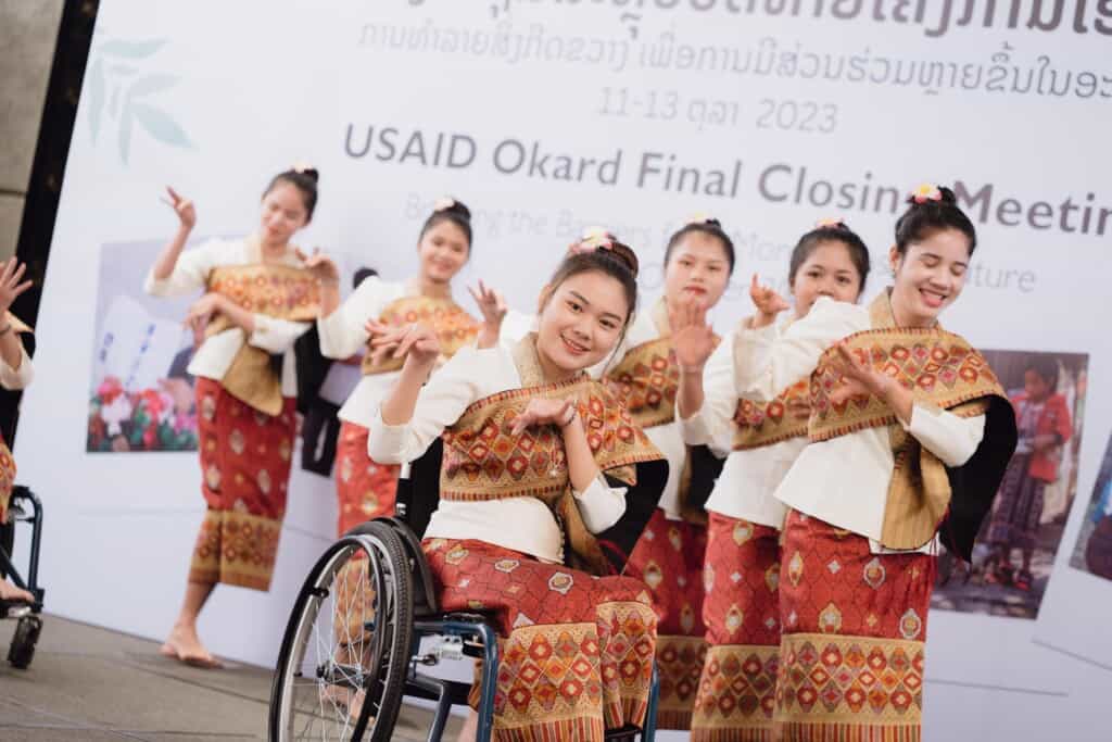 Group of Lao women, including a woman using a wheelchair, perform on stage at an event