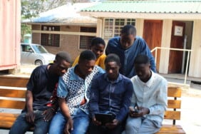 A group of men huddled around a man showing them how to use a phone