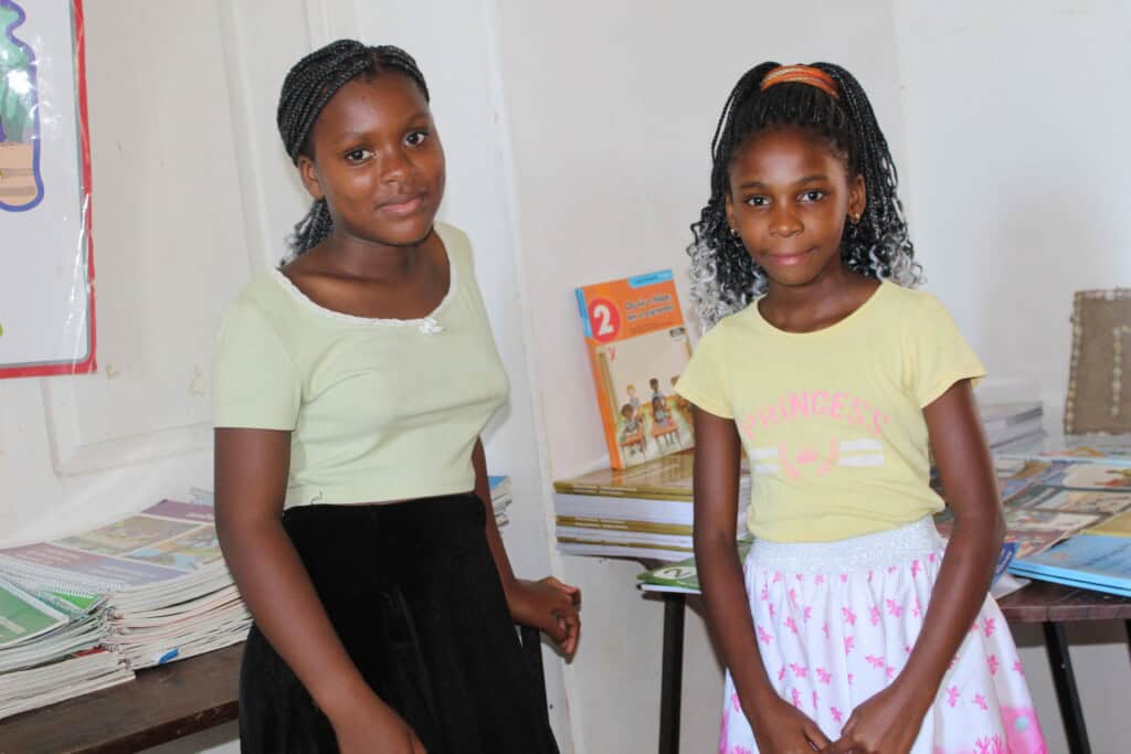 two young female students from Mozambique smile together in front of school books on tables