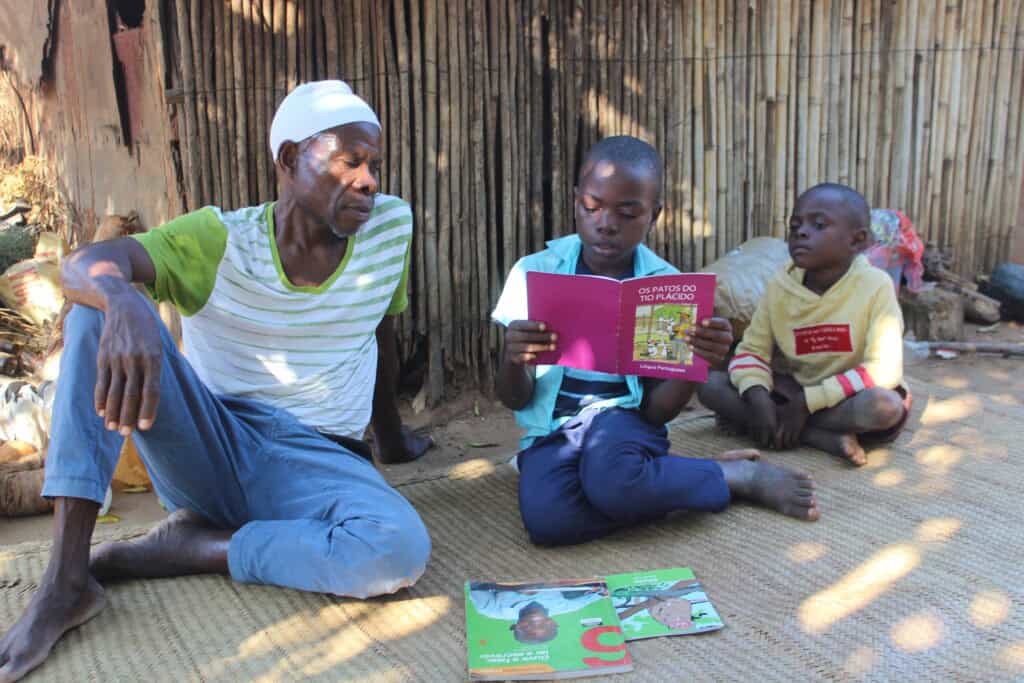 A man and his young sons sit outside their house while the older son reads a school book